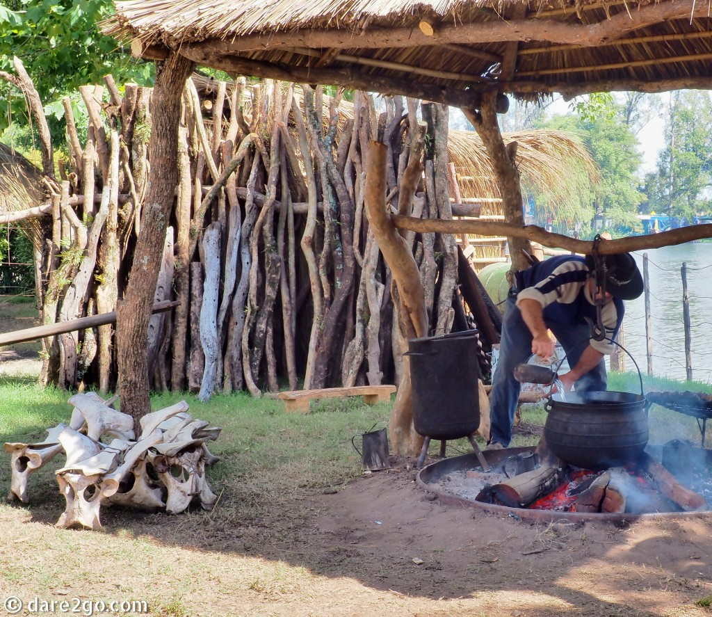 Calendar - May: typical gaucho cooking on the open fire. Above the fire hangs a cauldron to boil a meaty stew, next to the fire the meat is roasting on the 'asado' grill. Note the bone stools on the left!