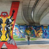 Under the overpass, looking back towards the retaining wall. In the foreground 3 columns with street art (and an unknown man).