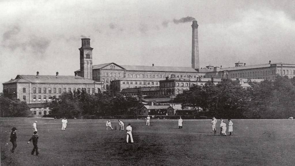 An historic photo of workers enjoying a game of cricket in the park on the other side of the Saltaire Mill and River Aire.