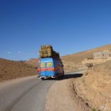 An old Mercedes Bremer as local transport. On the roof hay bales, some sheep in front of the hay, and passengers inside.
