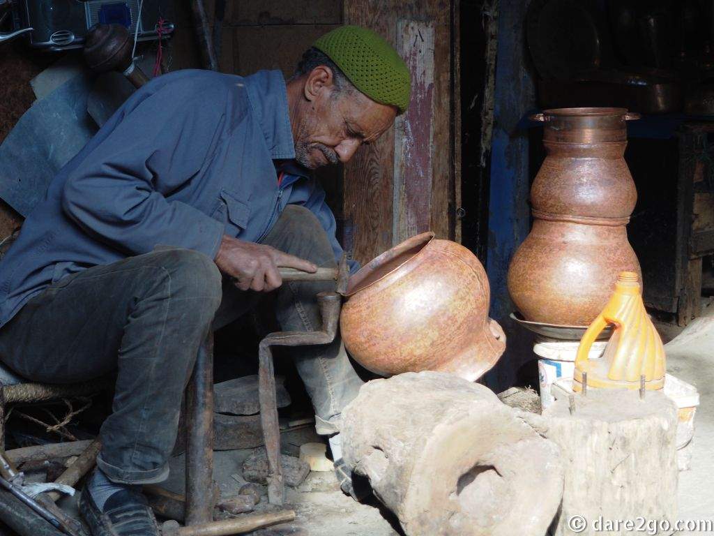 An old man sitting on a stool, hammering the rim of a copper pot into shape.