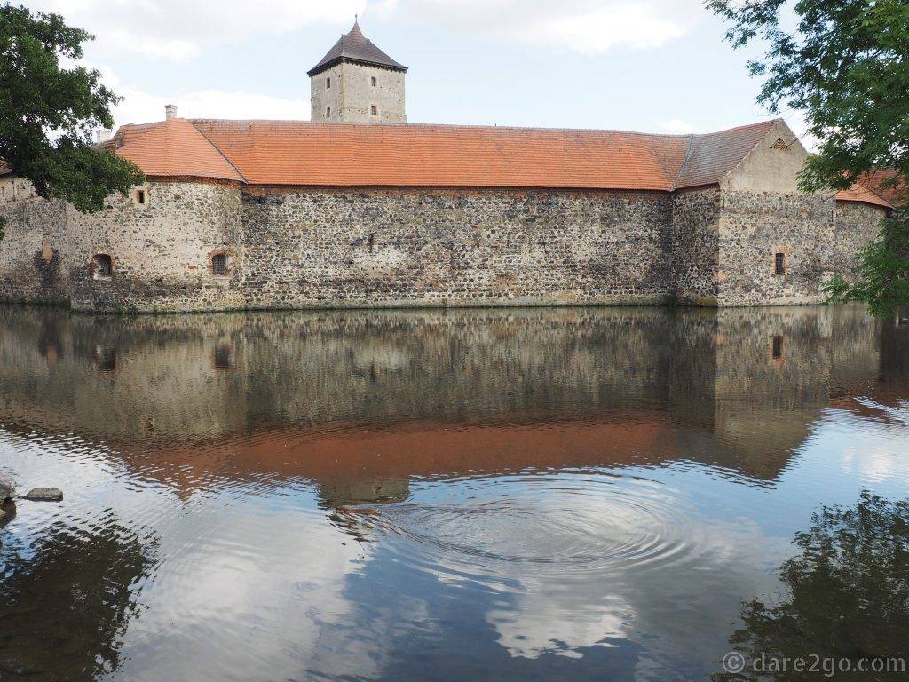 Panoramic view of the Svihov Water Castle, across its moat. There is a coypu/nutria swimming in the water.