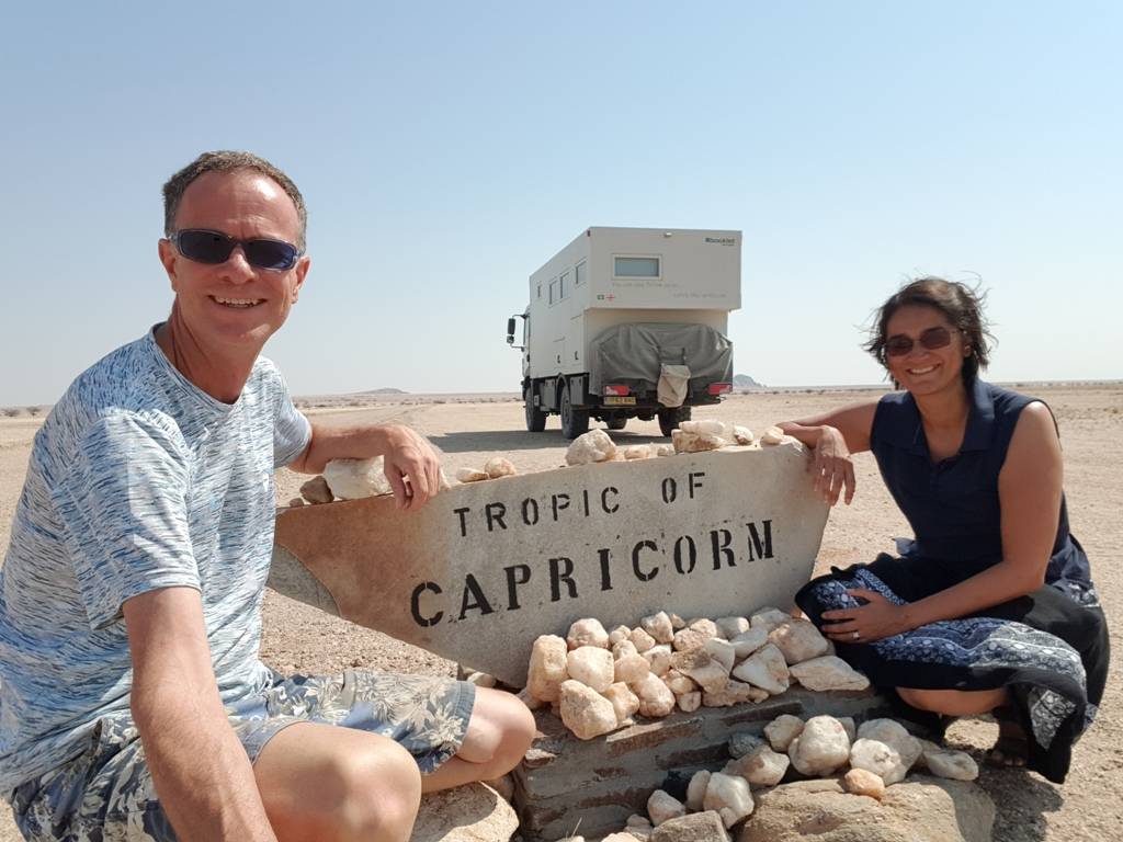 A milestone during our overland trip: crossing the Tropic of Capricorn [2 people squatting by a sign with a truck in the background]